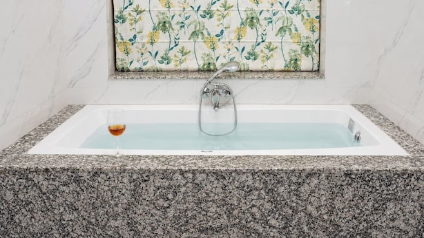 A bathroom with a rectangular bathtub, a shower, and a window with a floral pattern. - Kesar Bagh  (Ranakpur)
