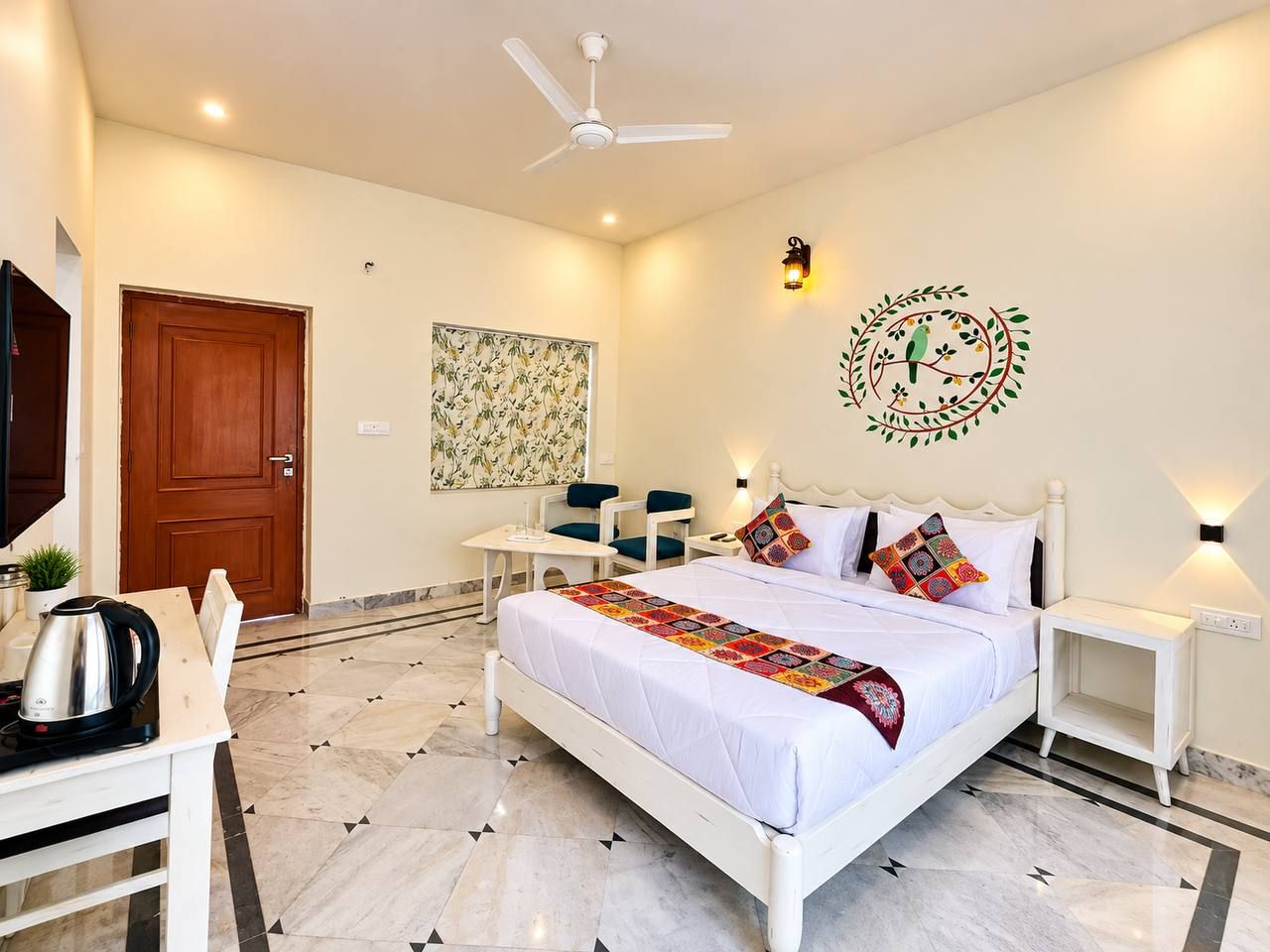 Quarto de hotel com cama, mesas de cabeceira, escrivaninha com chaleira e ventoinha no teto, além de luminária fixada na parede.