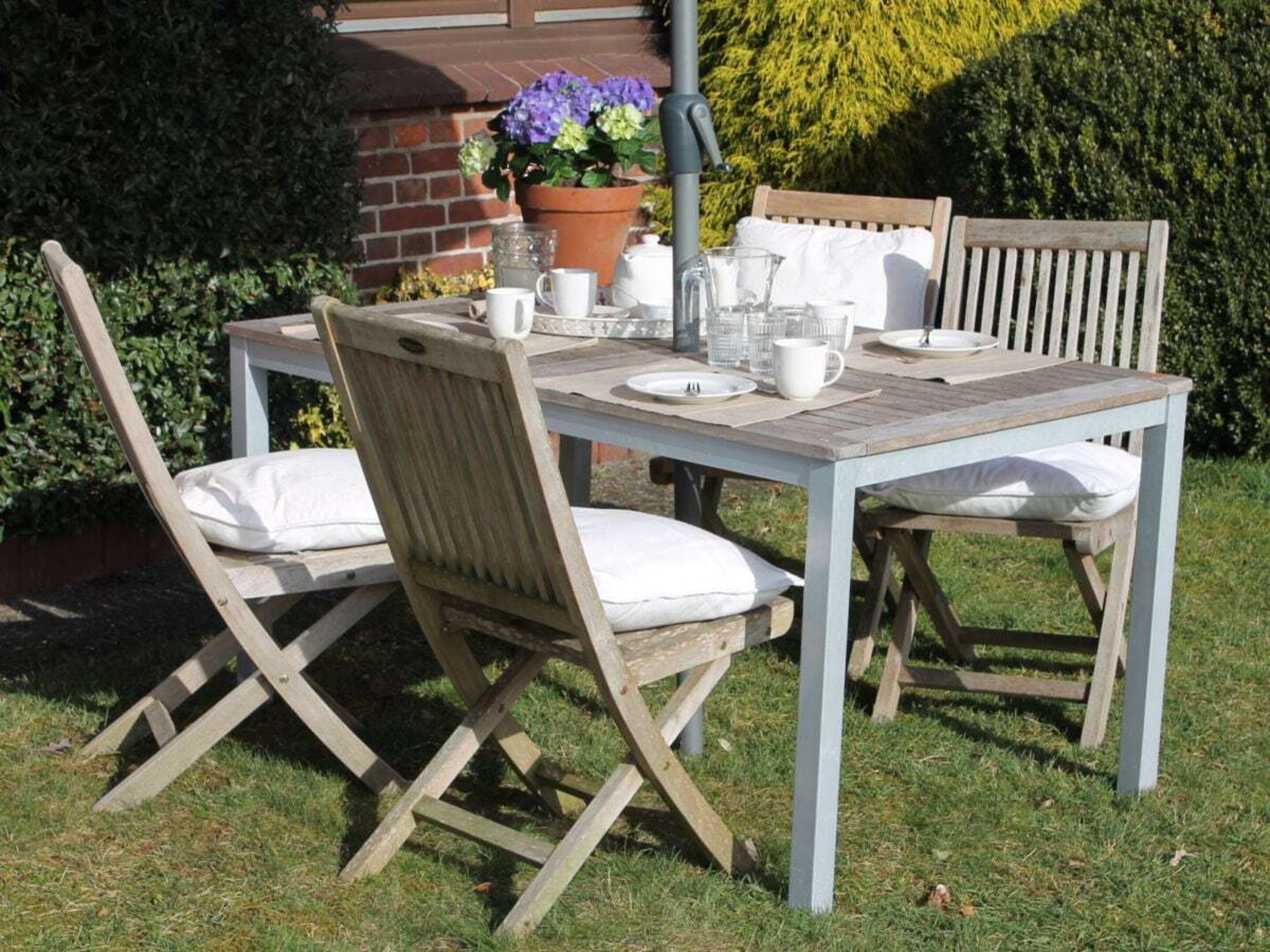 An outdoor wooden table set with white cushions, plates, and a potted plant.