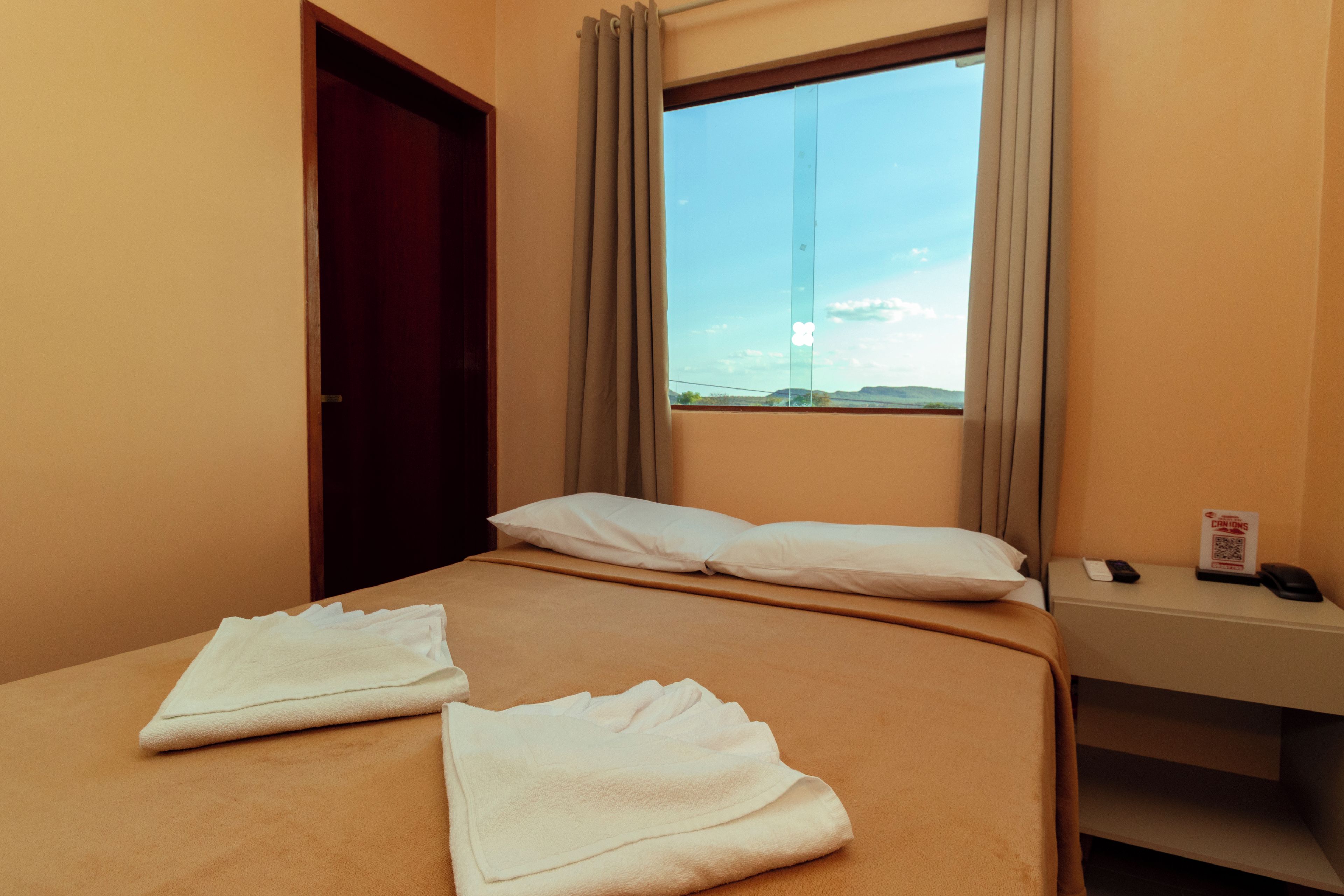Quarto de hotel com uma cama, janela com cortinas, mesa de cabeceira com um telefone e um sinal de código QR, e uma porta.