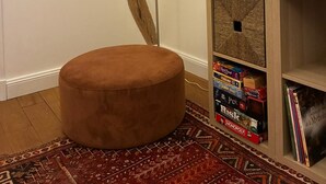 Een kamer met een plafond van houten balken, een rond poefje, een vloerlamp en een boekenkast met gezelschapsspellen.