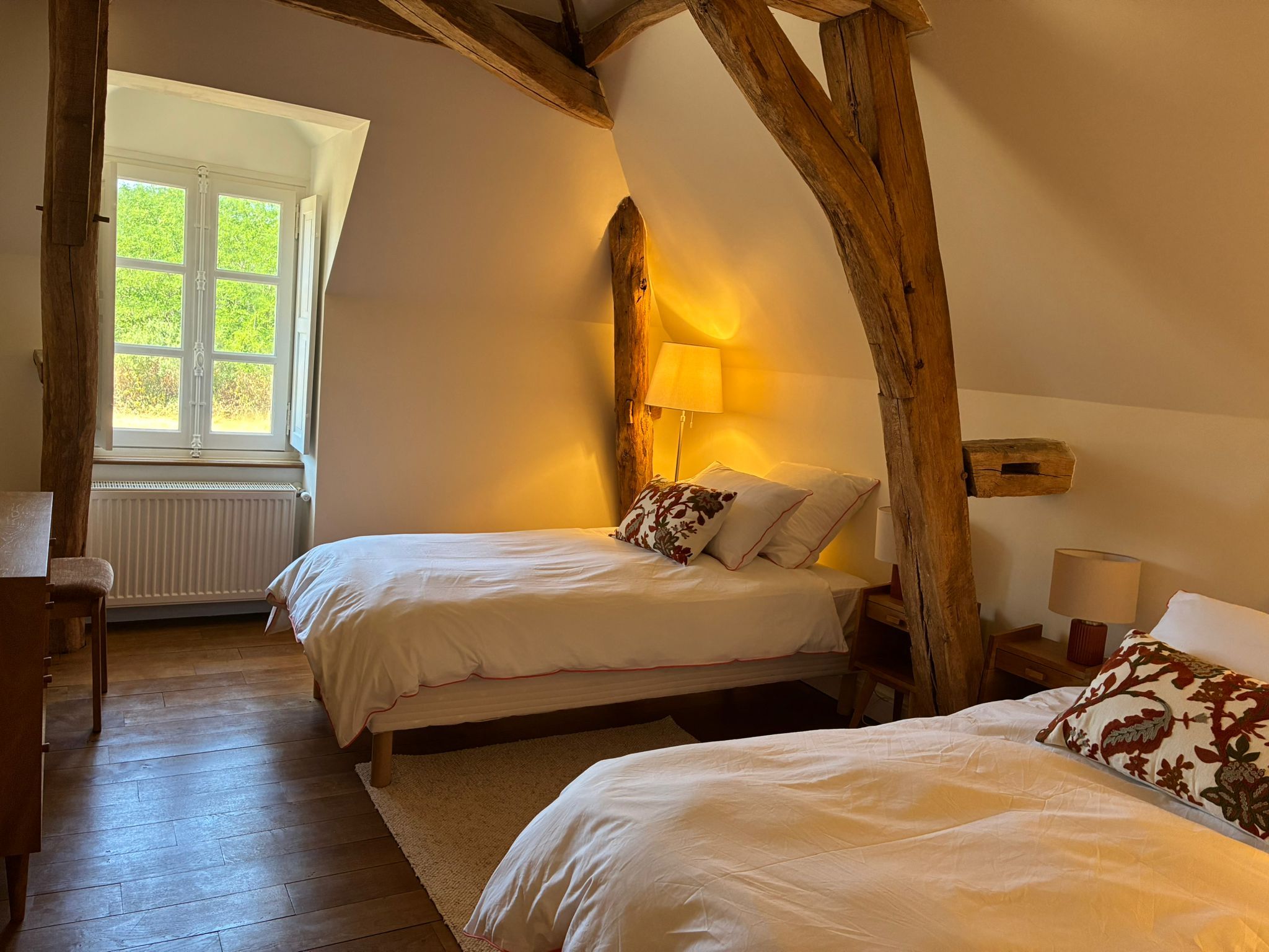 A room with two beds, a window, wooden beams, and a radiator.