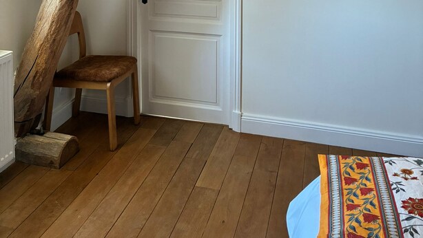 Een kamer met een schuine balkenlaag, een houten stoel, een witte deur en een tafelkleed met dessin.