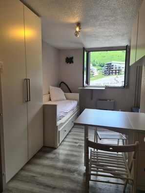 Habitación pequeña con cama, una mesita y ventana con vistas a edificios y vegetación.