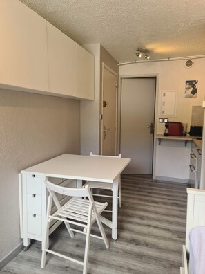 Ein kompakter Raum mit weißem Schreibtisch und Stuhl, einer kleinen Küchenzeile und einer Tür zu einem weiteren Raum.