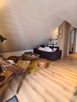 Een gezellige woonkamer met een houten vloer, een donkere bank, een houten bijzettafeltje en een klein rond voetenbankje.