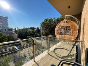 Un balcón con barandilla de vidrio, una silla de mimbre y vistas a edificios y árboles.