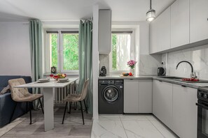 A modern kitchen with a built-in dishwasher, a dining area with a table and chairs, and a view through the windows. - Lumina 3 room apart near PGE Narodowy (Warszawa)