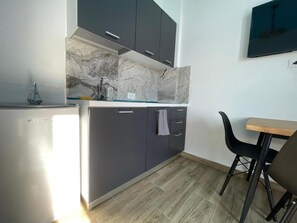 A compact kitchen with grey cabinets, a marble backsplash, a small refrigerator, and a dining area with a wooden table and black chairs. - Room-sea Pearl Studios Golem - Ground-fl (Golem)