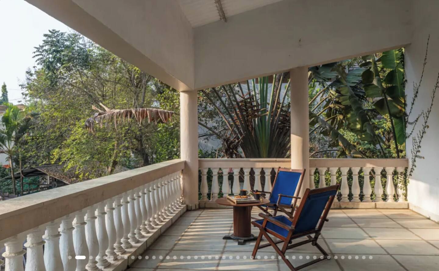 Ein überdachter Balkon mit Tisch und Stühlen, umgeben von einem weißen Geländer und üppiger Vegetation.
