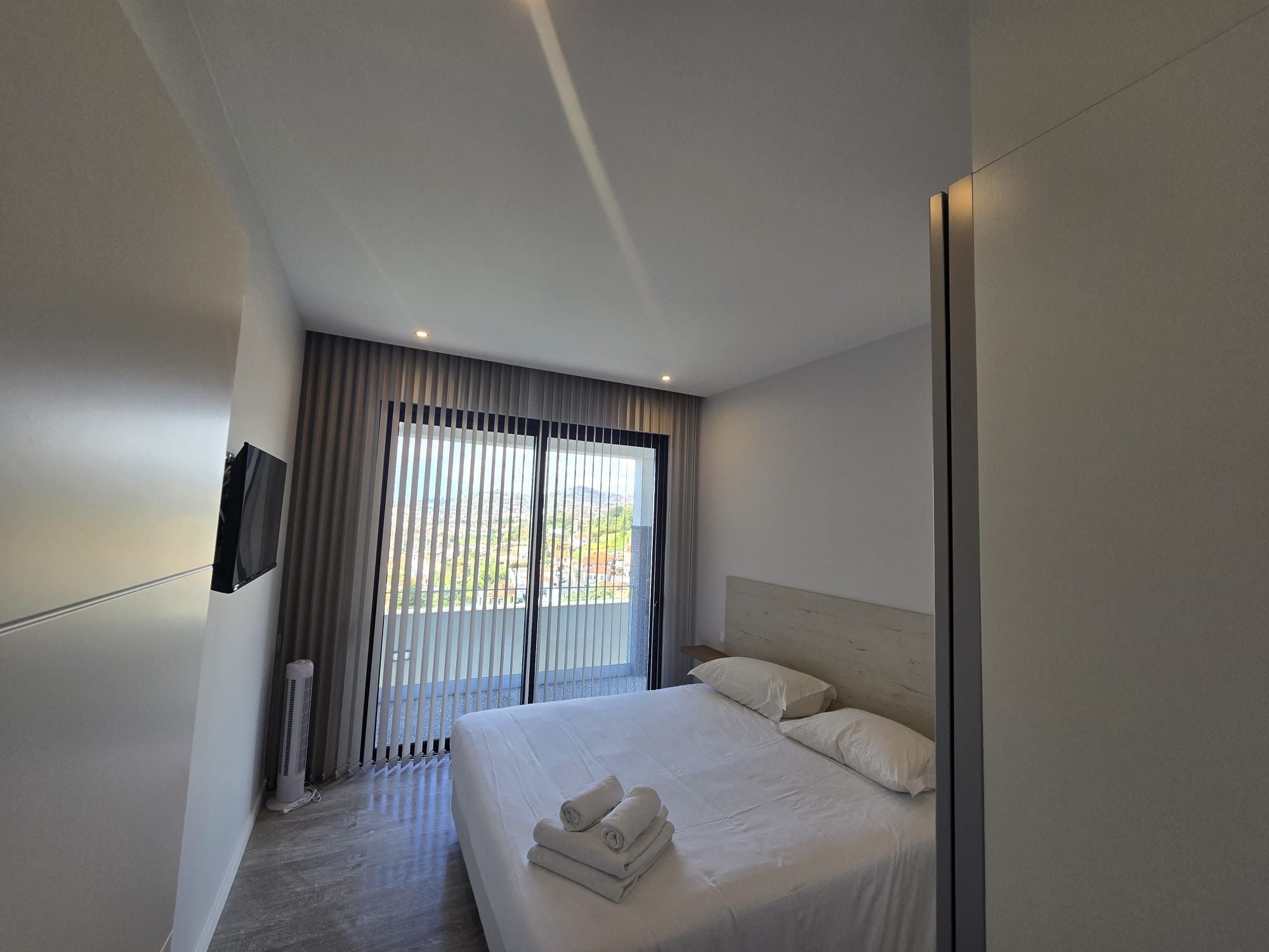A modern hotel room with a large bed, a flat-screen TV mounted on the wall, and a view of the outdoors through the window.