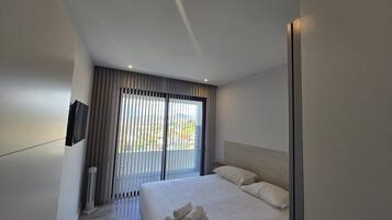 A modern hotel room with a large bed, a flat-screen TV mounted on the wall, and a view of the outdoors through the window.