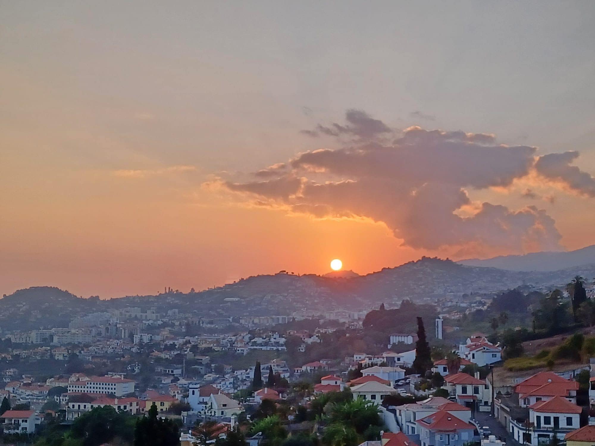 A cityscape with a sunset over a hill, featuring residential buildings with red roofs.
