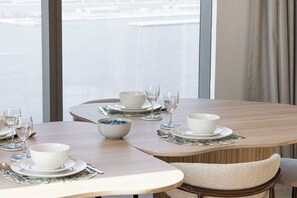 Une table à manger dressée avec des bols, des assiettes et des verres à vin blancs, offrant une vue sur une étendue d’eau grâce à de grandes fenêtres.