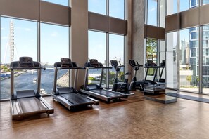 A gym with treadmills and elliptical machines, large windows, and a view of a river and buildings. - Ocean & Palm Views / Infinity pool / High floor (Dubai)