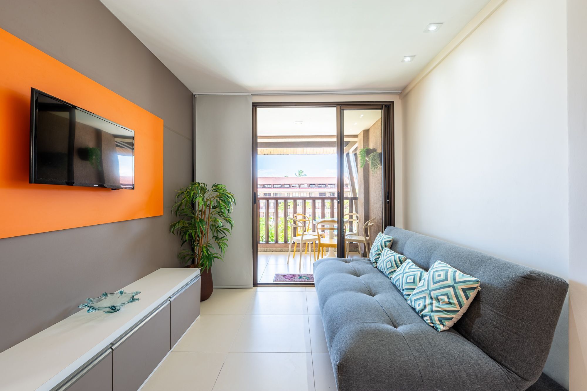 Un moderno salón con un sofá gris, un televisor de pantalla plana montado en una pared naranja, una planta en maceta y un balcón con mobiliario exterior.
