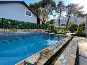 Una piscina con borde de piedra, rodeada por un seto y un edificio con ventana.