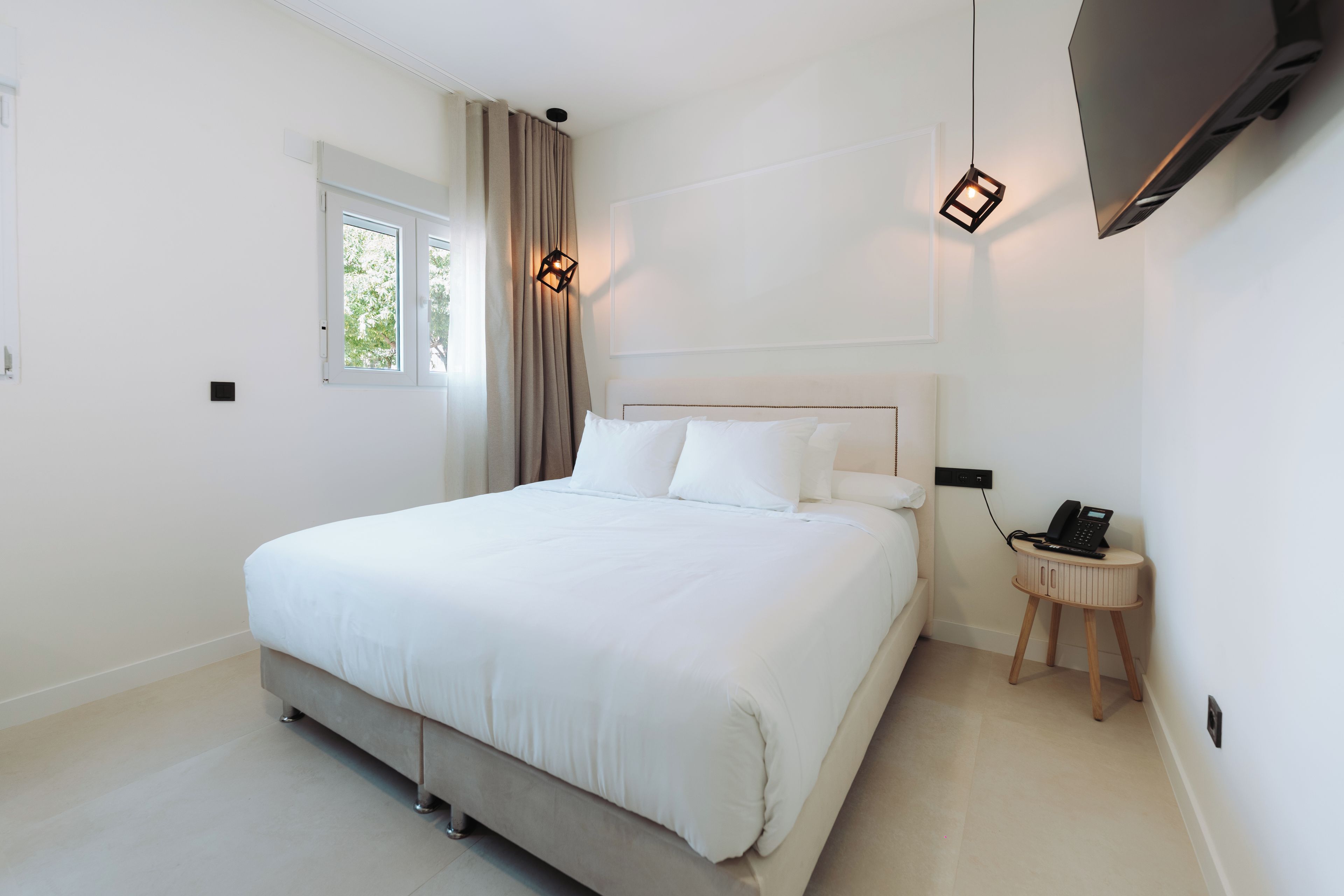 A modern bedroom with a large bed, a bedside table with a phone, and a window with curtains.