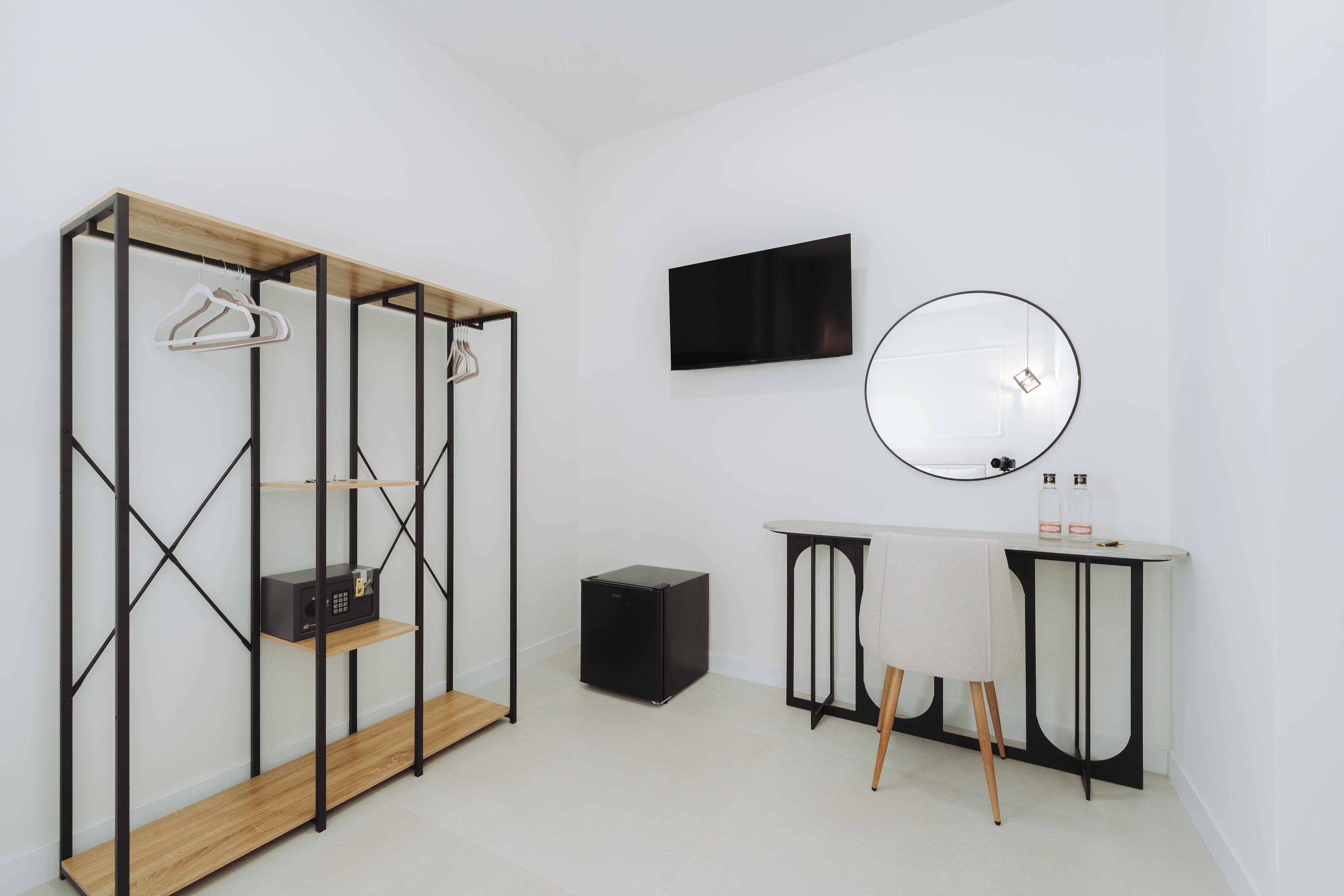 Une chambre moderne et minimaliste, dotée d’une armoire en bois, d’un téléviseur à écran plat, d’un miroir rond et d’une petite table sur laquelle sont posées des bouteilles.