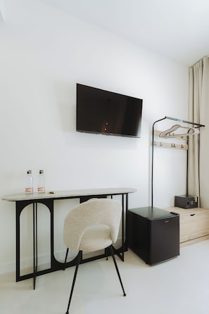 Una habitación moderna con un televisor de pantalla plana montado en la pared, un escritorio con dos botellas, una silla, un perchero y un altavoz negro.