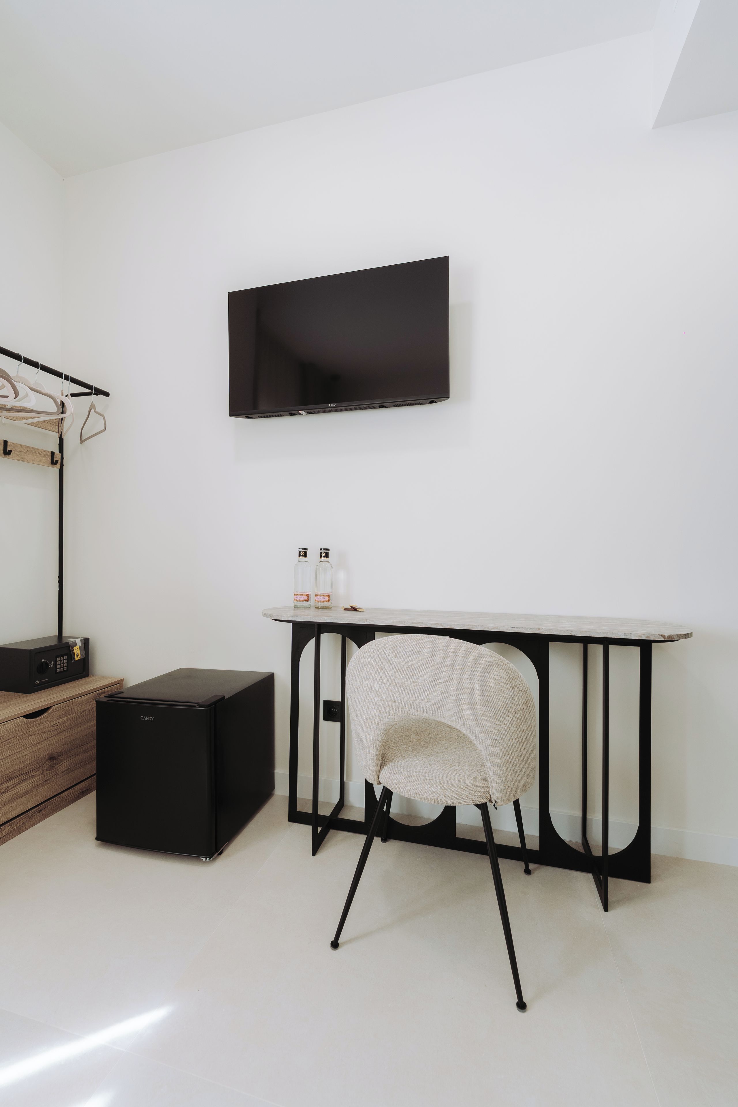 A minimalist room with a flat-screen TV mounted on the wall, a small round table with bottles, a chair, and a black storage unit.
