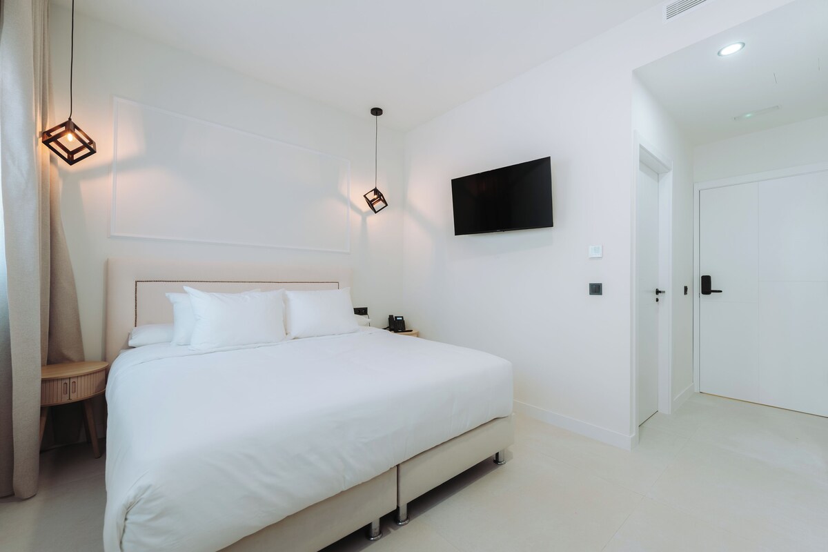 Habitación de hotel moderna con una cama grande, un televisor de pantalla plana en la pared y una puerta a la derecha.