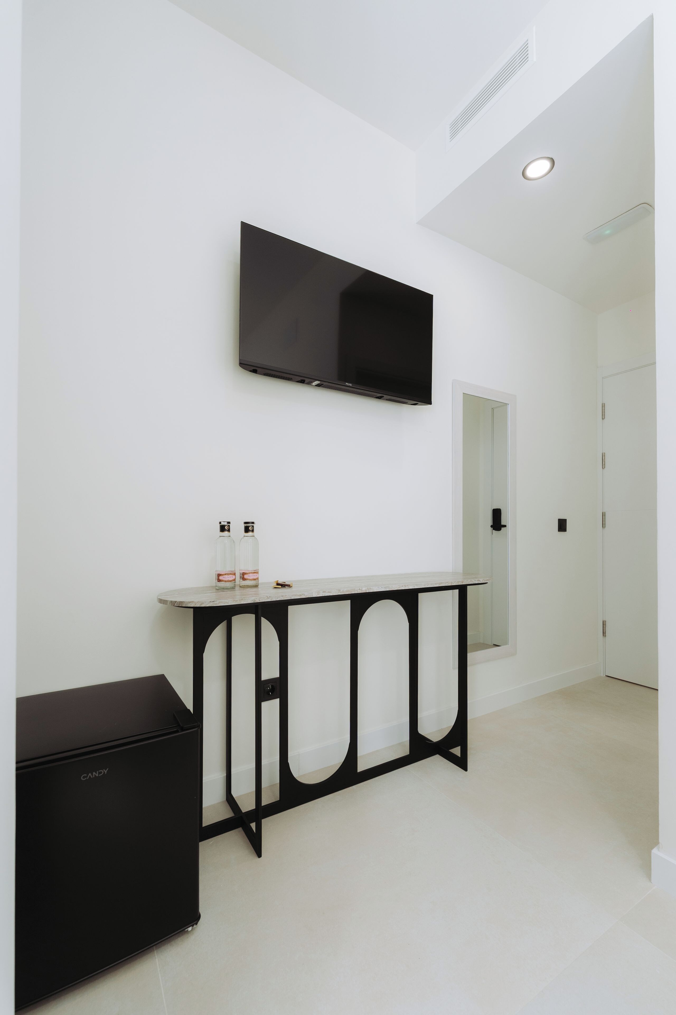 A modern, minimalist room with a flat-screen TV mounted on the wall, a small black cabinet, and a glass-fronted cabinet with bottles on a marble countertop.