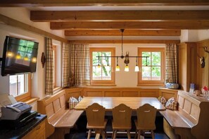 A wooden dining area with a large table and benches, a television mounted on the wall, and windows with curtains. - Das Jagdhaus im Großarltal (Großarl)