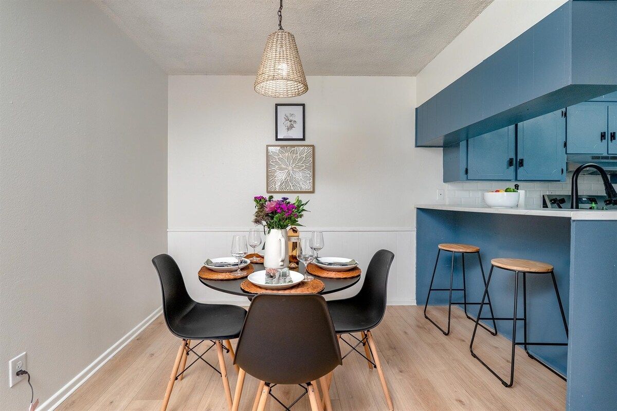 A dining area with a round table, black chairs, a hanging light fixture, and a kitchen with blue cabinets.
