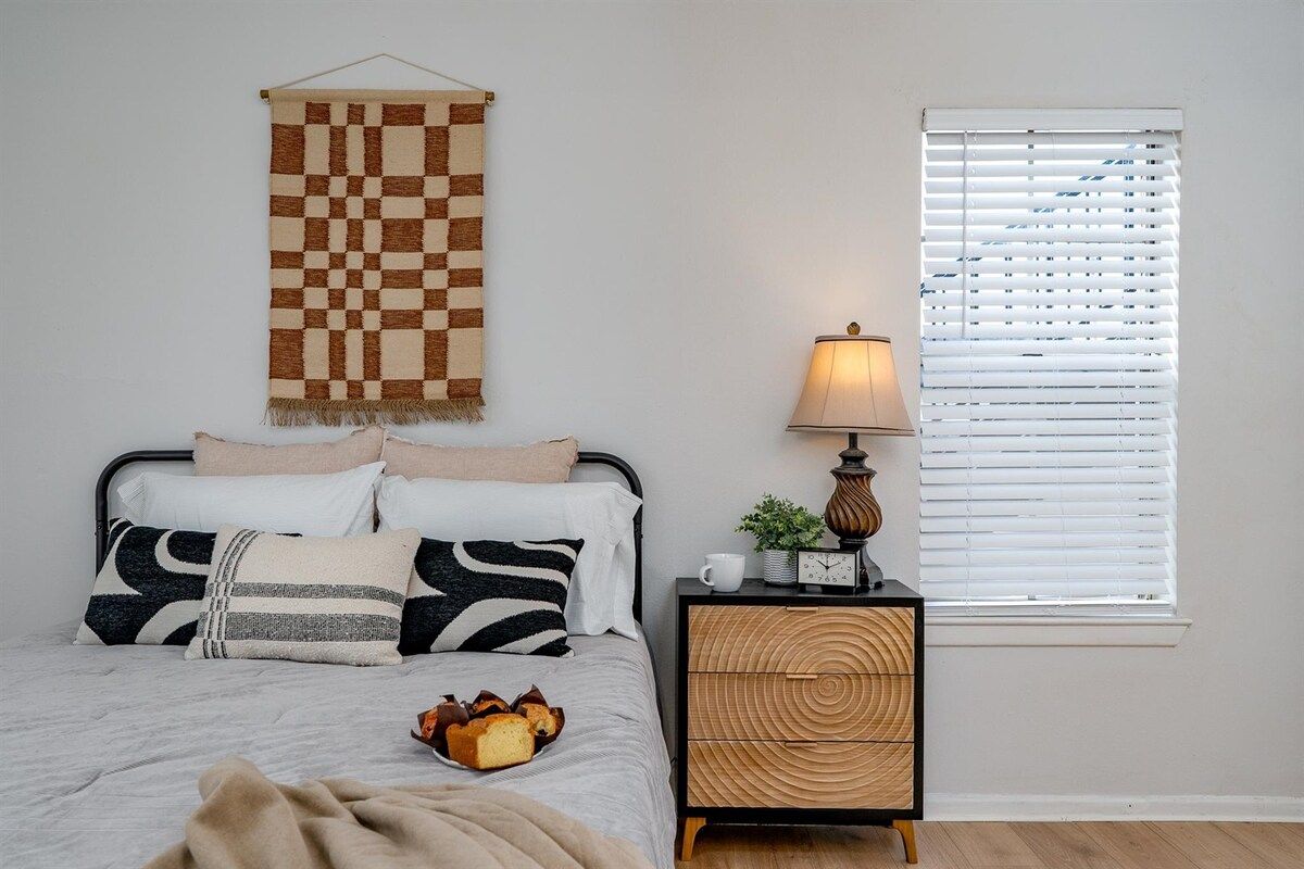 A bedroom with a bed, bedside table, lamp, and a window with blinds.