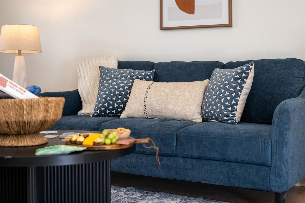 Ein dunkelblaues Sofa mit gemusterten Kissen, ein gewebter Beistelltisch mit Snacks und eine Lampe in einem Zimmer mit einem gerahmten abstrakten Kunstwerk an der Wand.