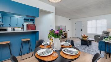 Eine moderne Küche mit blauen Schränken, ein Essbereich mit Tisch für vier Personen und ein Wohnzimmer mit Sofa und Lampe.