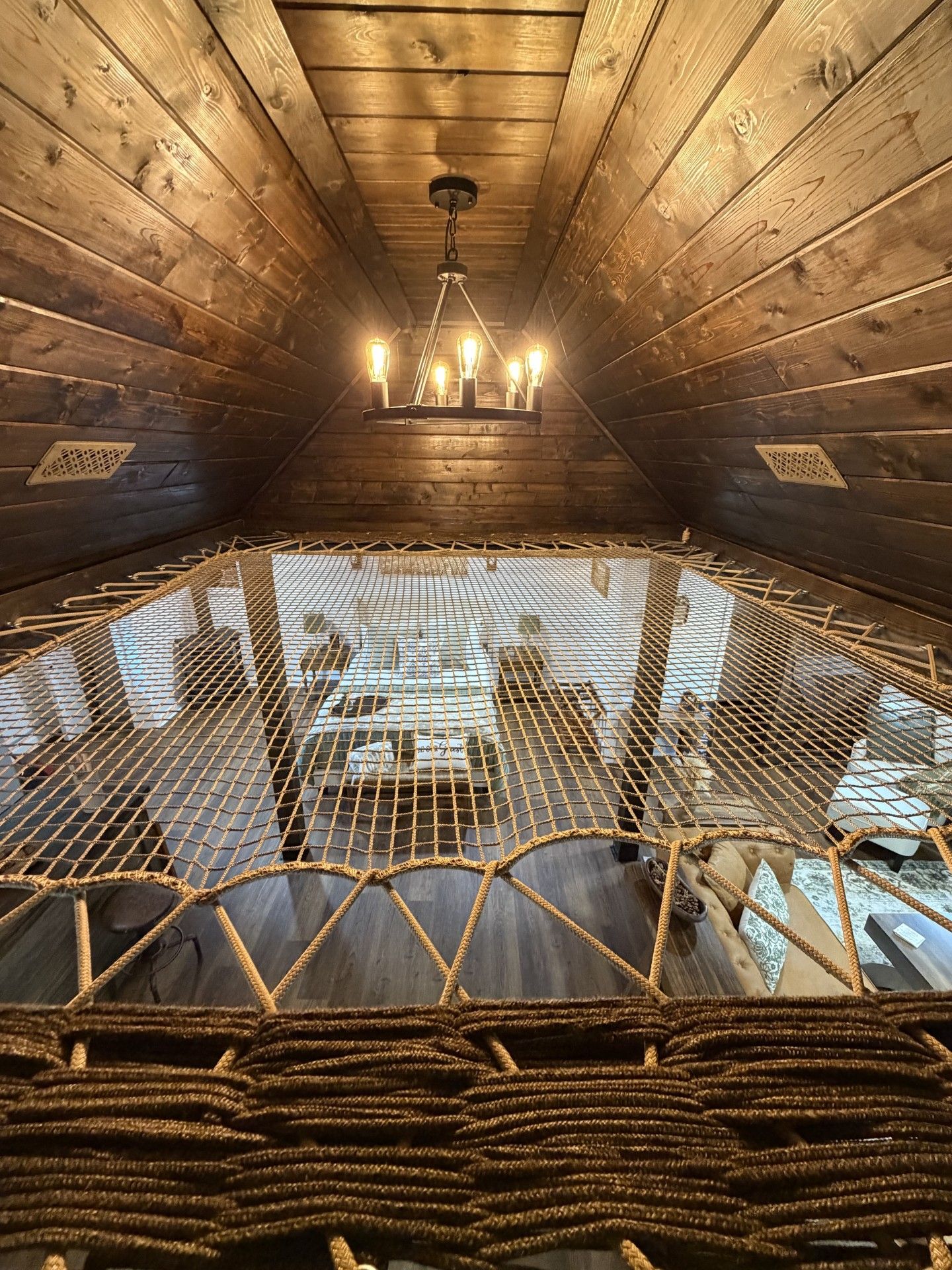A high-ceilinged room with wooden walls and a netted floor, featuring a chandelier and a view through the netting.