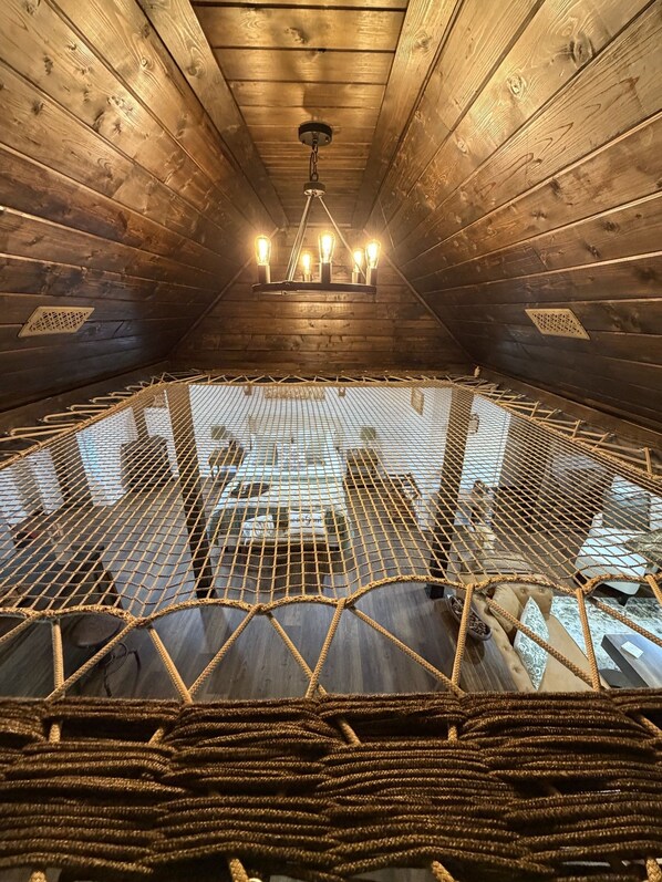 A high-ceilinged room with wooden walls and a netted floor, featuring a chandelier and a view through the netting.