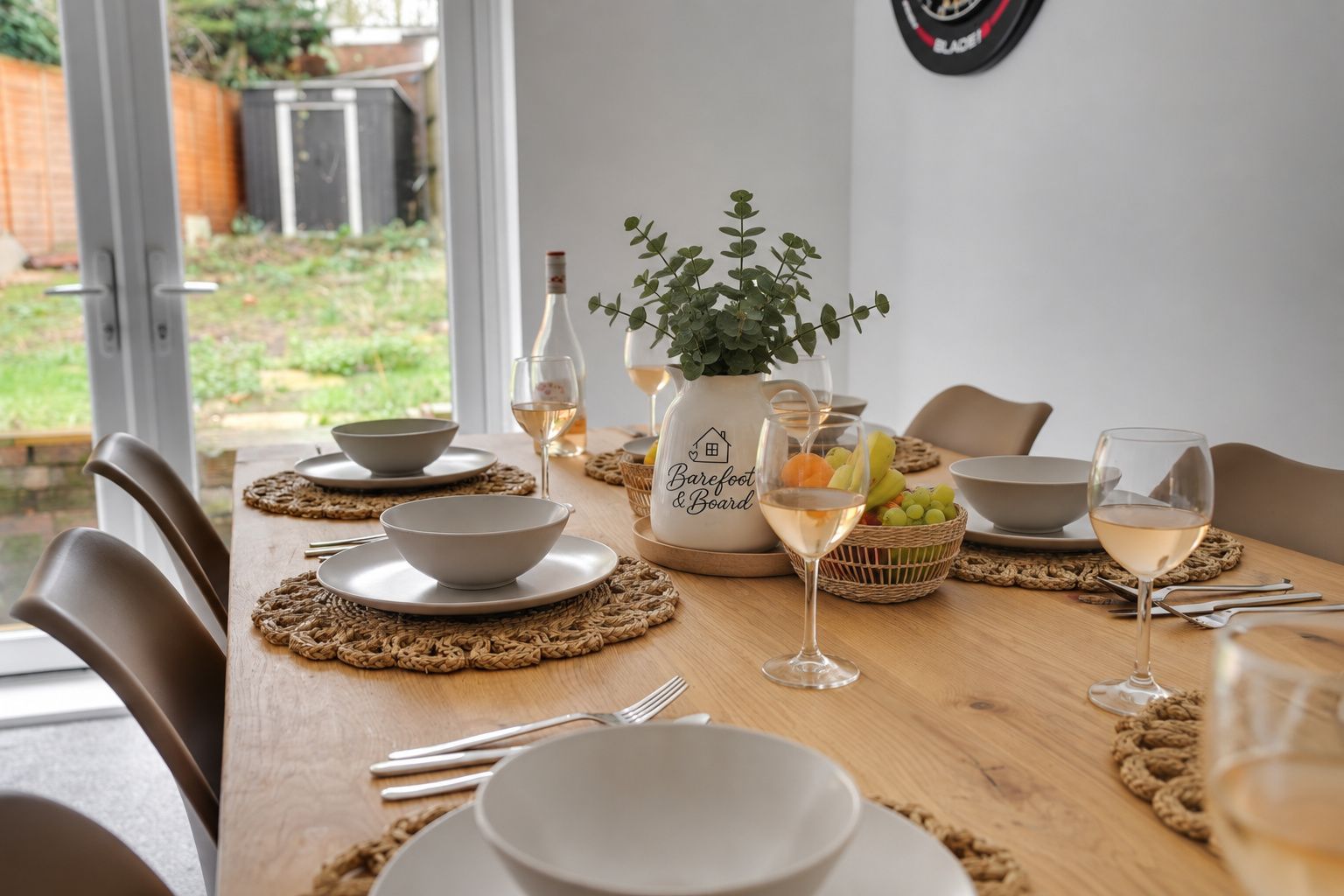 A dining table set with bowls, plates, cutlery, a pitcher, a glass of white wine, and a basket of fruit.
