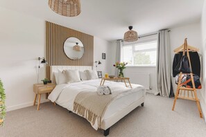 Un dormitorio con cama, mesitas de noche, lámpara de noche, espejo, lámpara colgante, ventana con cortinas y perchero.