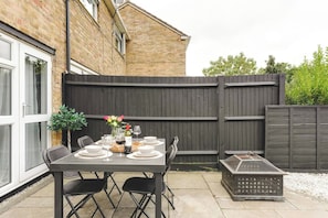 An outdoor patio with a table set for dining, surrounded by a wooden fence, and a brick building in the background. - Luxury 2-Bed House in Stevenage Sleeps 6 Free Parking Barefoot & Board (Shephall)