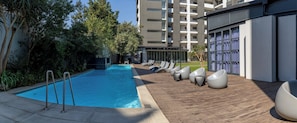 Ein Schwimmbecken mit Holzterrasse und moderner Sitzgelegenheit, umgeben von Grünflächen und einem mehrstöckigen Gebäude.