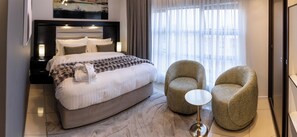 Ein modernes Hotelzimmer mit einem großen Bett, zwei Sesseln, einem runden Beistelltisch und einem Gemälde an der Wand.