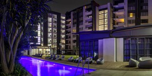 A modern hotel with a swimming pool, lounge chairs, and a well-lit building facade. - Bright 2-bedroom apartment with AC in Morningside (Sandton)