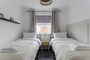 Ein Schlafzimmer mit zwei Betten, einem Nachttisch mit Lampe, einem Fenster mit Vorhängen und einem Regal mit Bildern.