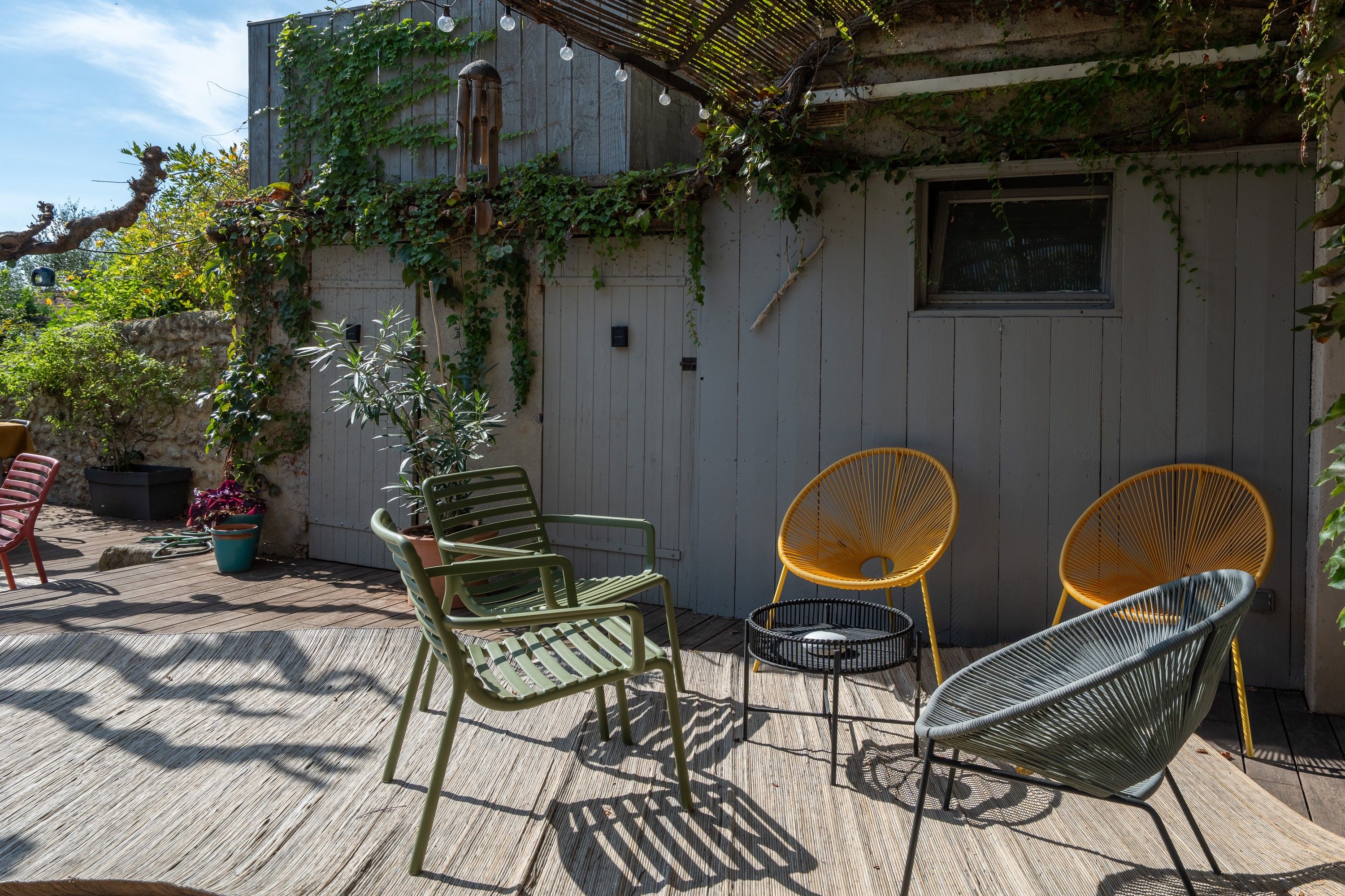 An outdoor patio with a wooden deck, a white wall with climbing plants, and two wicker chairs.