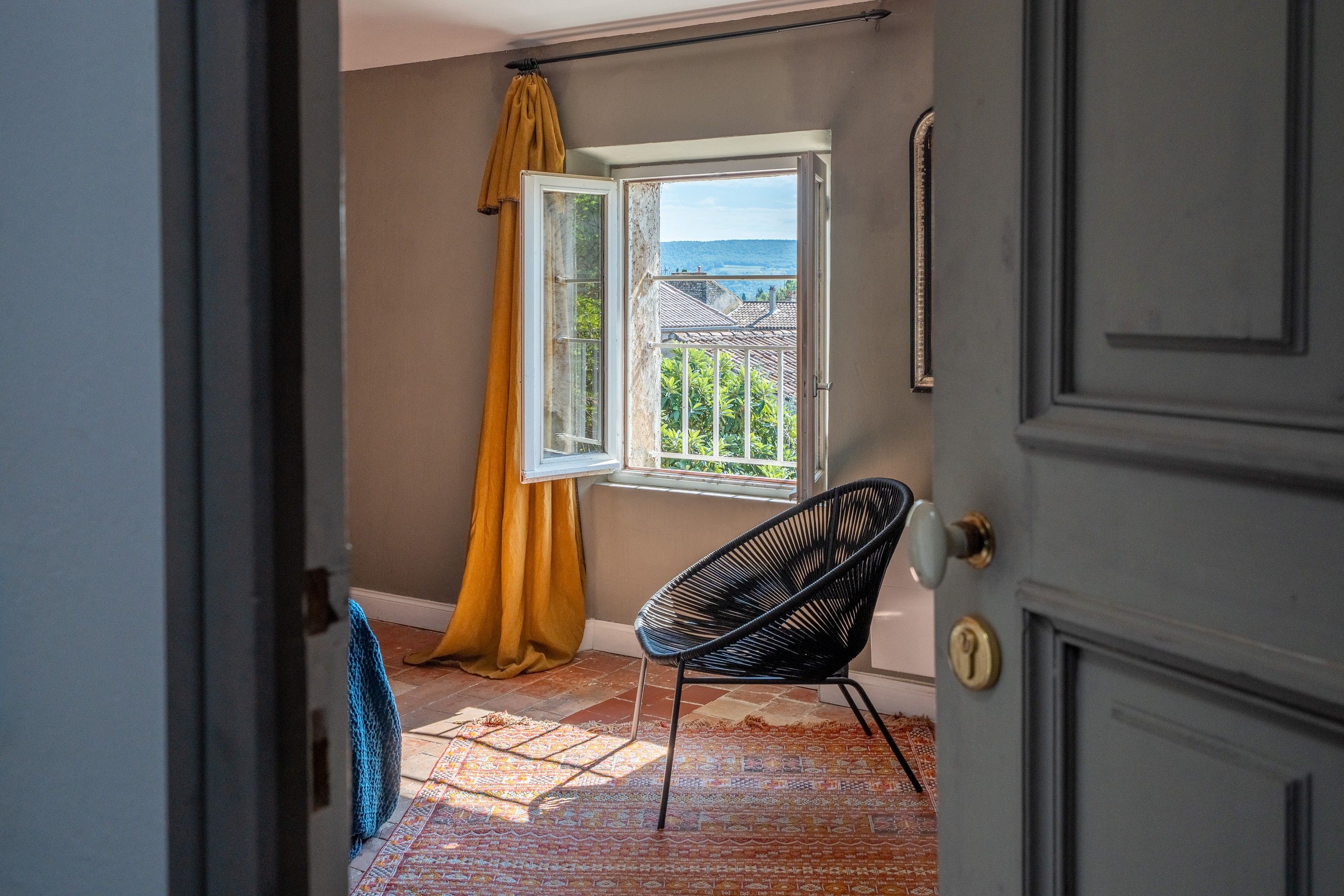 Une pièce avec vue sur une maison par une fenêtre ouverte, une chaise noire et un tapis à motifs.