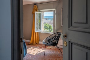 Una habitación con vistas a una casa a través de una ventana abierta, una silla negra y una alfombra estampada.