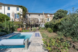 Una piscina con una tumbona y una mesa, rodeada de vegetación y un edificio con tejado de baldosas.
