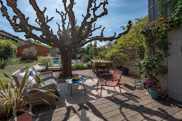 An outdoor patio with a table and chairs, surrounded by plants and a large tree.