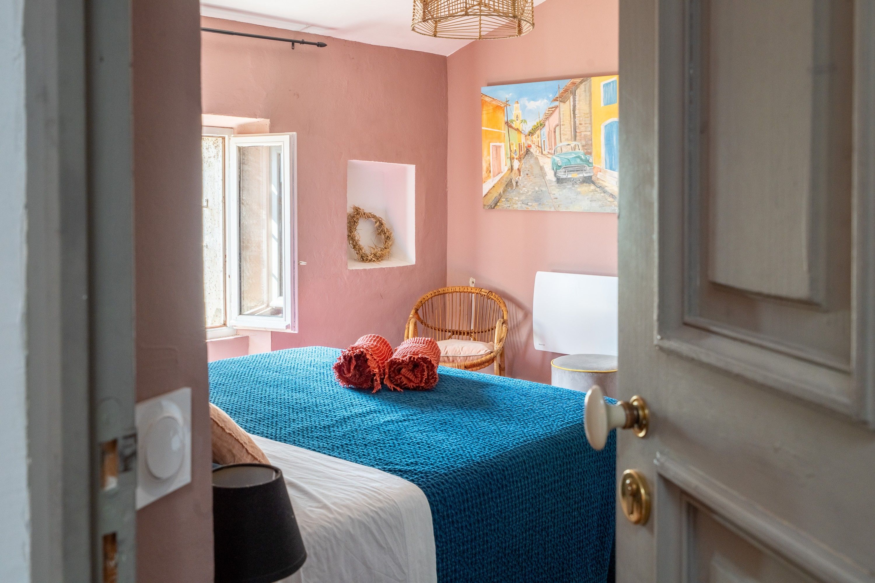 Une chambre à coucher avec un lit, une fenêtre, un tableau, une chaise en osier et une porte.