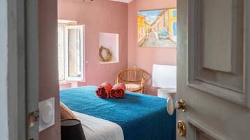 Une chambre à coucher avec un lit, une fenêtre, un tableau, une chaise en osier et une porte.