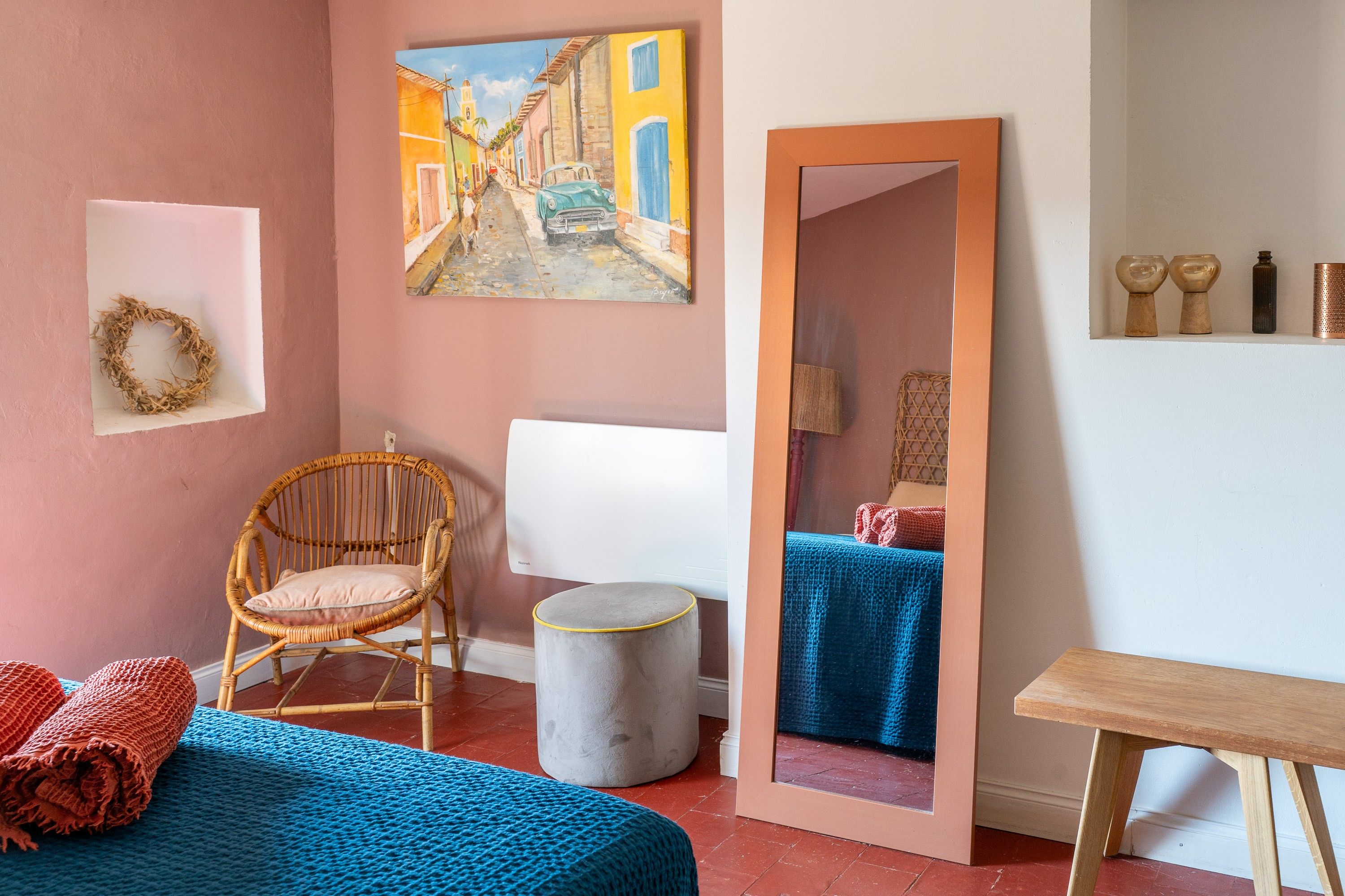 Una habitación con una cama, una silla de mimbre, un taburete pequeño, un espejo y un cuadro en la pared.