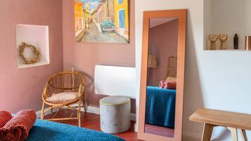 A room with a bed, a wicker chair, a small stool, a mirror, and a painting on the wall.